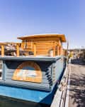 Blue houseboat with wooden deck and sign reading "Brighton Sunshine, Seattle, Washington" docked by a tree-lined path.