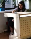Woman sitting at a wooden desk with pegboard design, near a red sofa and large potted plants by a window.