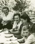Family gathered around an outdoor table, smiling and serving soup, with trees and a wooden fence in the background.