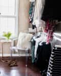 Walk-in closet with hanging clothes, clear chair with fur pillow, small table, and leopard print heels on wooden floor.