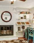 Living room with white brick fireplace, round mirror, teal desk, and built-in shelves with books and decor.