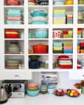 Open kitchen shelves with colorful vintage bowls, plates, and cookware, alongside a kettle and toaster on the counter.