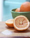 Halved grapefruit on wooden board with salt, green bowl of whole grapefruits in background.