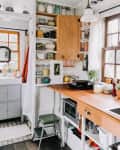 Cozy kitchen with wooden countertops, potted plants on windowsill, open shelves with mugs, and a sink with black faucet.