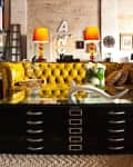 Loft living room with a yellow tufted sofa, glass coffee table, and vibrant orange lamps on a textured rug.