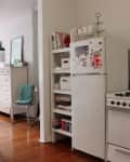 Small kitchen with white fridge, stove, and open shelving. Fridge has magnets, and shelves hold books and baskets.