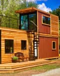 Two-story tiny house with wood siding, spiral staircase, and rooftop deck, set in a wooded area.