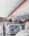 Airplane cabin with passengers seated, watching screens on seatbacks, and a flight attendant standing near the exit.