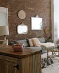 Loft-style living room with brick wall, gray sofa, wicker room divider, and green bedspread.