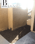 Small outdoor area with beige stucco walls, mulch ground, and partial wooden fence under bright sunlight.