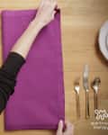 Folding a purple napkin on a wooden table with gold cutlery, a white plate, and a vase of red berries.
