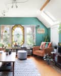 Cozy living room with teal walls, beige sofa, orange armchair, plants, and a stained glass window.