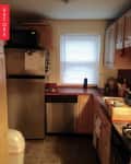 Small kitchen with stainless steel fridge, dishwasher, wooden cabinets, red countertops, and a window with blinds.