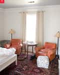 Bedroom with two orange leather armchairs, a round wooden table, patterned rug, and beige curtains.