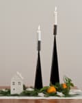 Two black candle holders with lit candles, a small white house, greenery, and oranges on a table runner.
