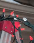 Person on ladder hanging red and white Christmas lights outdoors.
