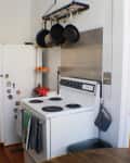Small kitchen with white fridge, vintage stove, hanging pans, spice rack, and view into a bedroom with wooden floors.