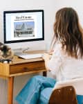 Woman working at a wooden desk with a computer displaying a real estate listing, cat resting beside her.