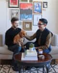 Two people sitting on a sofa with a dog, surrounded by eclectic wall art, books, and a candle on a coffee table.