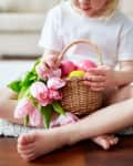 Child sitting on a rug holding a basket with pink tulips and colorful eggs.