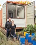 Couple standing in front of a shipping container home with a garden of blue planters.