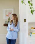 Woman in blue shirt holding a mug, standing in a kitchen with fruit bowl and hanging plant.