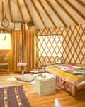Cozy yurt interior with colorful textiles, wooden furniture, and a patterned rug on hardwood floors.