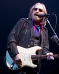 Musician with long hair and sunglasses playing an electric guitar on stage, wearing a dark jacket and colorful scarf.