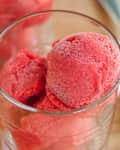 Bright red sorbet scoops in a clear glass, with a wooden surface and a spoon in the background.