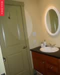 Small bathroom with beige walls, a round mirror, wooden vanity, and a sink with soap dispensers.