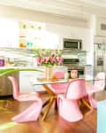 Bright kitchen and living area with pink chairs, glass table, blue sofa, tropical cushions, and neon "Paradise" sign.