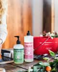 Woman washing hands at kitchen sink with Mrs. Meyer's hand and dish soap, surrounded by flowers and a red bucket.