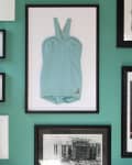 Framed vintage blue swimsuit on teal wall with black and white photos and small decorative plates.