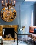 Elegant living room with blue walls, gold mirror, chandelier, and patterned armchair near a glass table and fireplace.
