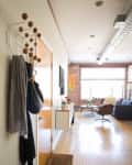 Hallway with wooden floors, coat rack, and a view into a living room with a sofa, armchair, and large window.