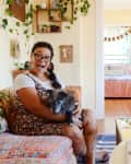 Woman with glasses holding a small dog on a colorful sofa in a cozy living room with plants and wall art.