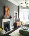 Living room with gray walls, colorful artwork, white fireplace, green ottoman, and wooden dresser near a large window.