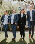 Groomsmen in blue suits and white vests walking on grass, surrounded by greenery and trees.