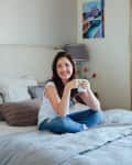 Woman sitting on a bed with a mug, wearing a white shirt and jeans, with a painting and lamp in the background.