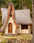 Fairy-tale cottage with a curved shingle roof, wooden door, and stone path surrounded by lush greenery.