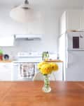 Kitchen with white cabinets, wooden table, yellow flowers in a vase, and a hanging light fixture.