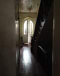 Dimly lit hallway with wooden floor, staircase, and arched doorway leading to a bright entrance.