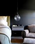 Bedroom with a hanging globe light, gray floating nightstand, and brown armchair next to a bed with white linens.