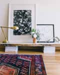 Floral artwork and abstract line drawing on a low wooden shelf with a brass lamp, vase of flowers, and decorative antler.