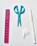 Blue scissors, pink ruler, white hooks, and colorful kitchen utensils in a tray on a countertop.
