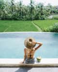 Woman in straw hat sitting by an infinity pool with a coconut drink, overlooking lush green rice fields and palm trees.