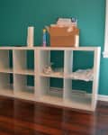 White cube storage unit against teal wall, with cardboard box and cleaning supplies on top.