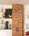 Open kitchen shelves with white ceramics, colorful vases, a red pot, and decorative rooster figurines beside a brick column.