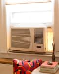 Window air conditioner in a cozy room with colorful cushion, books, and potted plants on a wooden shelf.
