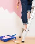 Person painting wall pink while standing on ladder, paint tray and roller on floor.